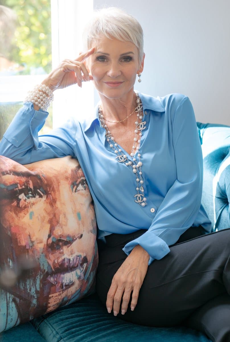 Linda in a blue blouse and pearls, seated on a teal sofa, chin on her hand
