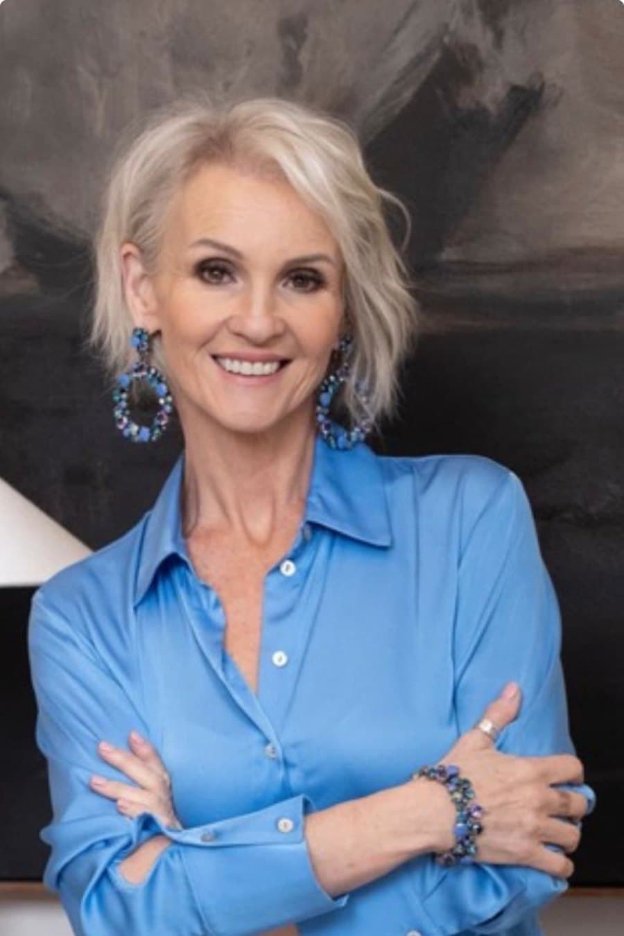 Linda Paige in a blue silk shirt with sapphire statement earrings, soft wavy bob, photographed against a charcoal painterly backdrop