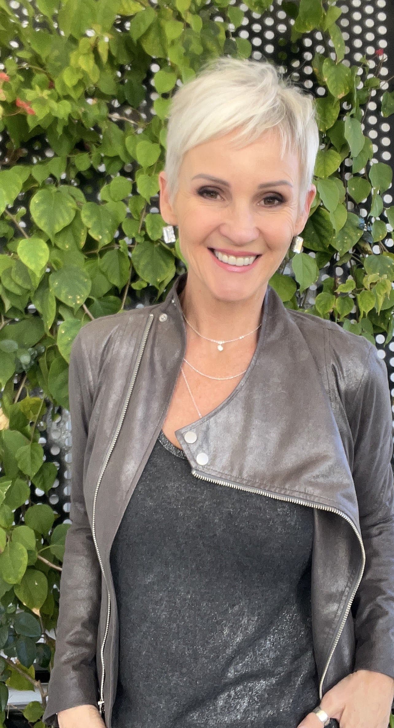 Linda smiling in a grey top and a grey leather jacket in front of green leaves