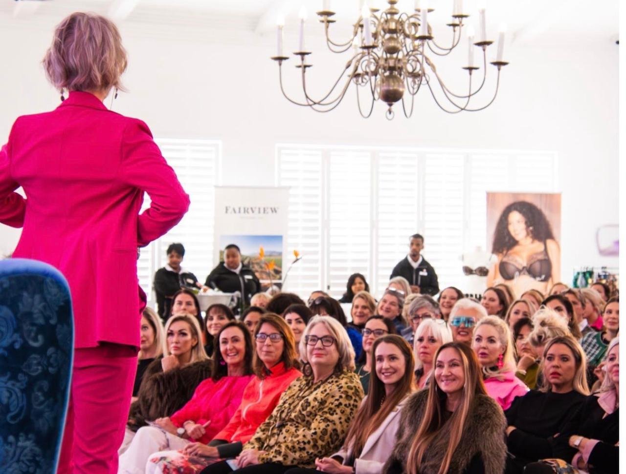 Linda Paige on stage in a pink suit, speaking to a packed room at one of her live events